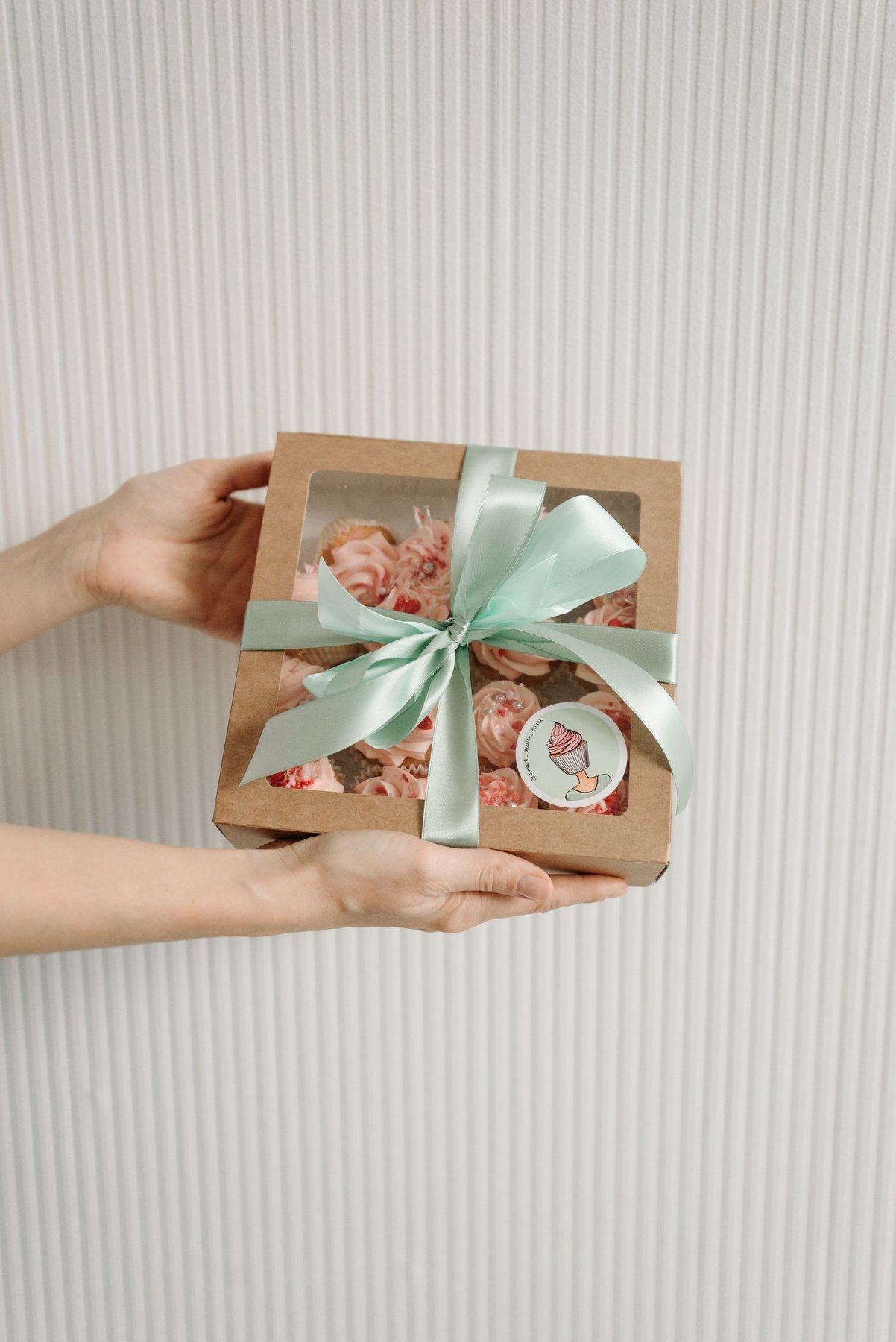 A person holding a windowed cupcake box finished with a pale ribbon