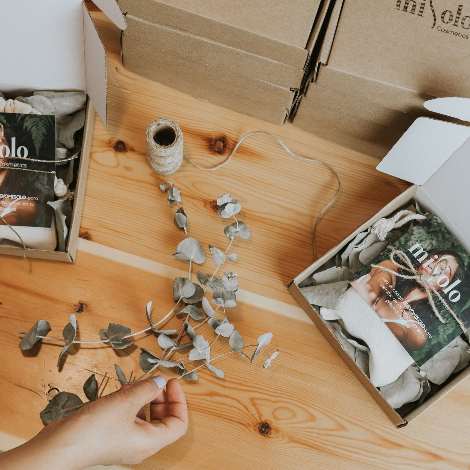 Beauty products being prepared with paper boxes and eucalyptus leaves at a worktable