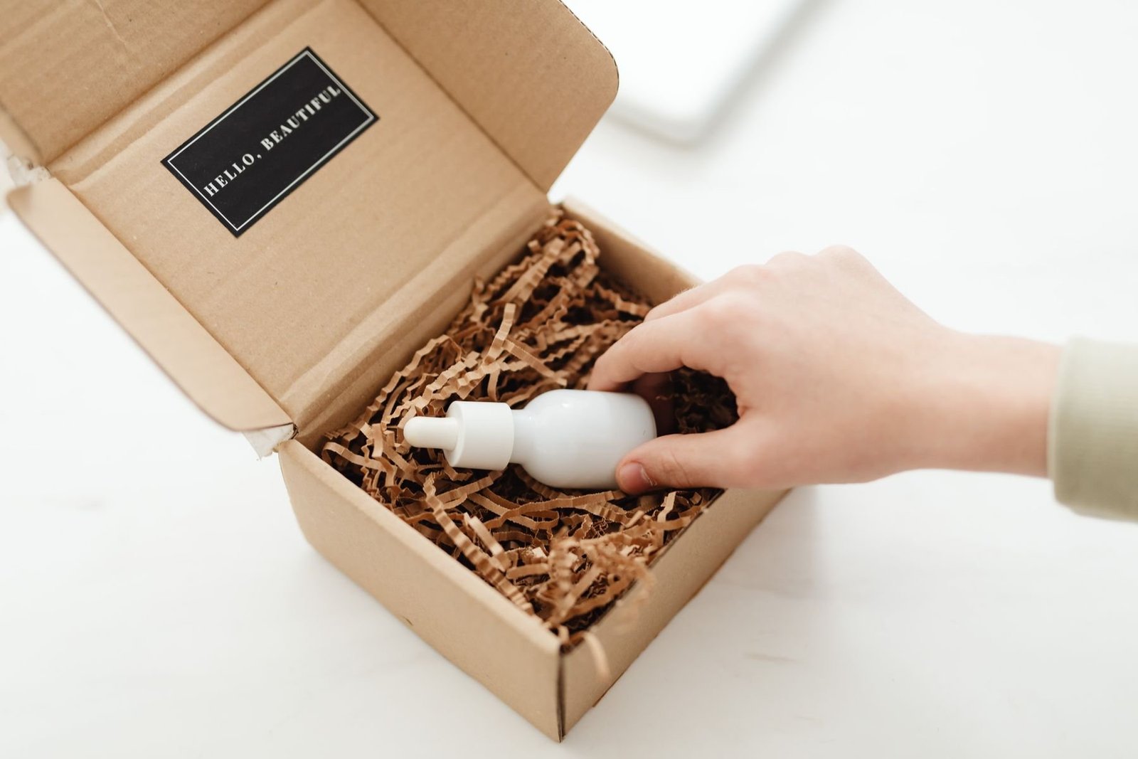 A hand lifting a white beauty bottle from a cardboard box with shredded paper