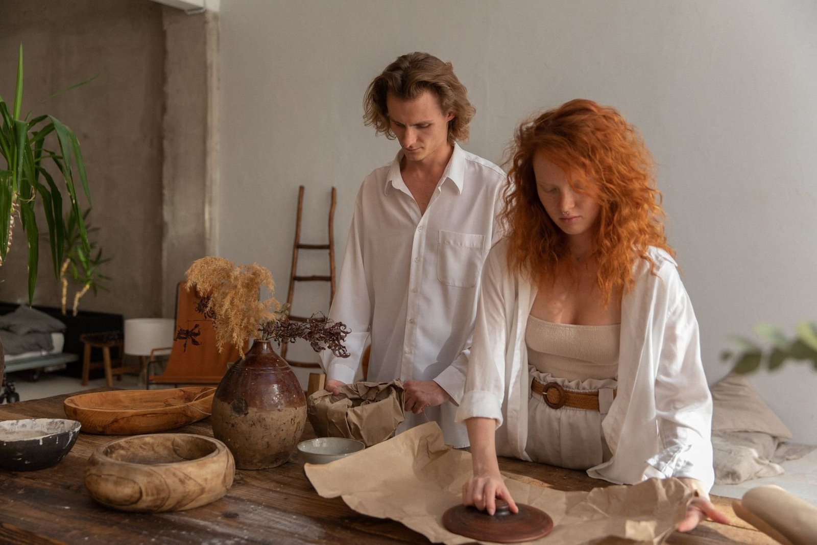 Two people wrapping ceramic bowls in brown paper at a workshop table